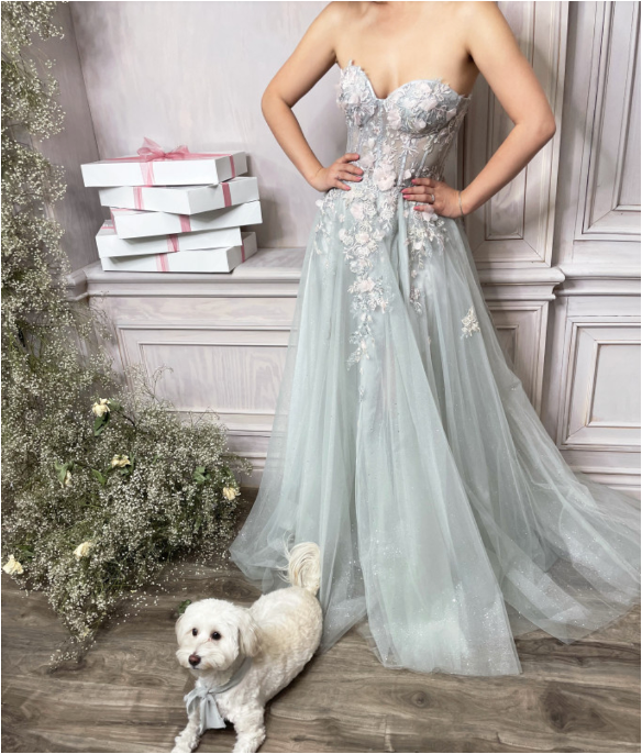 Strapless ball gown by Andrea & Leo, style A1089. Features intricate and delicate details with sheer floral appliqué corset bodice, leg slit, and over skirt. Available in Sage, Paris-Blue, and Blush-Mauve colors.