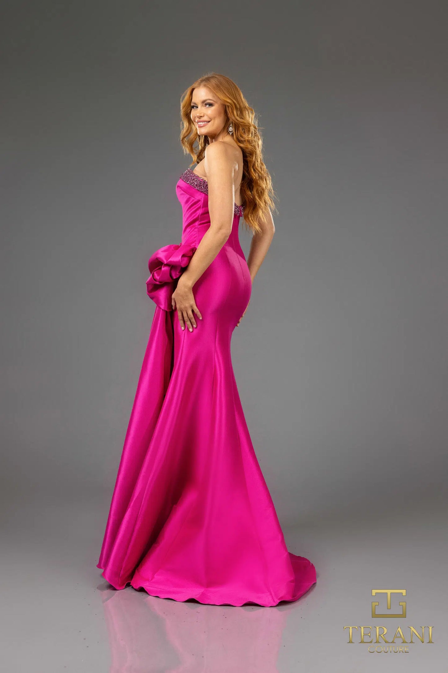 Terani 252E5131 strapless gown with embellished bodice, draped bow detail, and side slit, perfect for formal evenings and mother of the bride or groom.  The model is wearing the dress in the color fuchsia.